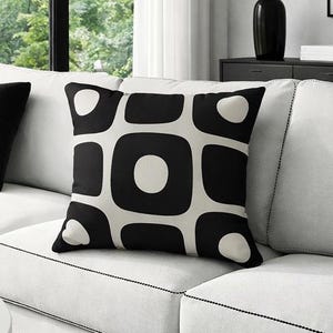 Puede incluir: Salón moderno con sofá blanco, cojines negros y un cojín decorativo estampado. Una mesa de centro redonda exhibe una taza negra, gafas y un libro con la palabra "DESIGN". Una lámpara de pie y un jarrón completan la decoración.