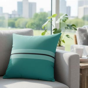 Op de afbeelding: Een turquoise decoratief kussen met een horizontaal ontwerp van twee turquoise strepen en een zwarte streep. Het kussen ligt op een grijze bank voor een raam met uitzicht op de stad. Een salontafel met een kopje en glazen staat naast de bank.