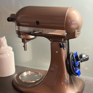 Könnte beinhalten: Ein bronzefarbener Artisan-Küchenmixer mit poliertem Finish. Der Mixer hat einen abgerundeten Kopf und einen stabilen Sockel mit einem Aufsatz für eine transparente Glasschüssel. Das Netzkabel ist ordentlich aufgewickelt und mit blauen Clips befestigt. Der Text "Made in USA" ist sichtbar.
