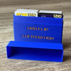 Könnte beinhalten: Ein blauer 3D-gedruckter Kartenhalter mit dem Text "UNO FLIP!" und "LUCA EDITION". Der Halter enthält zwei Stapel UNO-Karten. Eine separate, passende blaue Ablage befindet sich vor dem Halter.