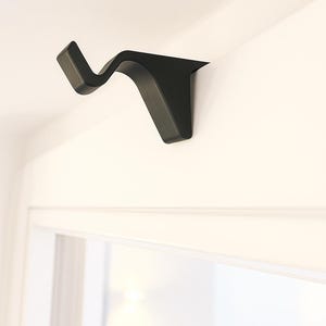Könnte beinhalten: Ein schwarzer Gardinenstangenhalter, der an einer weißen Wand befestigt ist. Der Halter hat ein unverwechselbares, geschwungenes Design in Wellenform. Er befindet sich über einem Fensterrahmen. Der Halter besteht aus einem soliden Material.