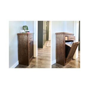 Op de afbeelding: Een houten kast met een donkerbruine afwerking, met een lade en een scharnierende deur. De deur is op één afbeelding open en onthult een vuilnisbak binnenin. De kast staat tegen een muur in een gang.