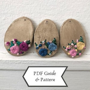 Puede incluir: Tres adornos beige en forma ovalada con diseños florales bordados. Cada adorno presenta una combinación de colores diferente: rosa y morado, azul y amarillo. El texto "PDF Guide & Pattern" se muestra en una pancarta blanca debajo.