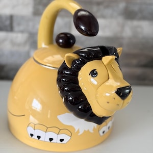 May include: A yellow tea kettle shaped like a lion, featuring a dark brown mane, a sculpted lion head spout, a matching dark brown handle, and a dark brown lid knob. The body of the kettle is decorated with white and black graphic details.