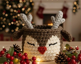May include: A handcrafted crochet reindeer basket in brown, cream, and grey. It has a red nose, closed eyes, and grey antlers. Red berries, pine cones, and fairy lights decorate the scene. A Christmas tree and fireplace are visible in the background, creating a festive atmosphere.