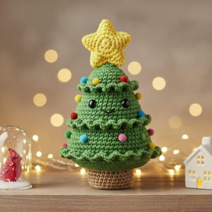 Pode incluir: Uma árvore de Natal de crochê verde artesanal com um rosto sorridente, adornada com pompons coloridos e uma estrela amarela. A árvore fica em uma base de crochê marrom, criando uma decoração de férias festiva e caprichosa.