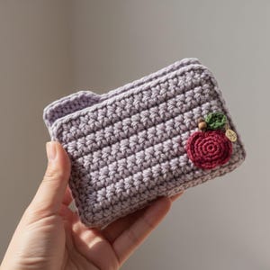 May include: A crocheted wallet in a soft lavender hue, featuring a textured stitch pattern. A red apple applique with green leaves and a small gold charm adorns the front. The wallet is held in a hand, showcasing its handmade quality.