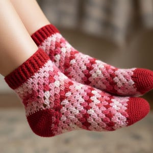 May include: A pair of handmade crochet socks in shades of red, pink, and white. The socks feature a textured, granny square pattern with solid red cuffs and toes. The socks are designed for comfort and warmth.