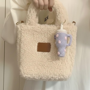 Puede incluir: Bolso tote cremoso y esponjoso con un parche rectangular marrón. Un llavero de ganchillo con forma de taza de bebida morada clara con lunares rosas cuelga de la bolsa. La bolsa tiene un asa corta.