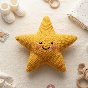 Könnte beinhalten: Ein gelbes, gehäkeltes sternförmiges Babyspielzeug mit einem lächelnden Gesicht und rosa Wangen. Der Stern ist von anderen Babyartikeln umgeben, darunter weiße Babyschuhe, ein Holzbeißring und eine Decke mit goldenen Sternen.