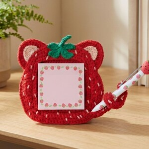 Pode incluir: Um porta-memorandos em forma de morango vermelho de crochê com um bloco de notas rosa em branco e uma caneta a condizer. O porta-memorandos tem uma folha verde e detalhes de costura brancos. A caneta tem um topo de morango e um corpo branco com padrões de morango.
