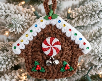 Puede incluir: Adorno navideño de casa de jengibre de ganchillo marrón con ribete de glaseado blanco y detalles de gemas de colores. Un caramelo de menta poivrée rojo y blanco está centrado en la casa, con dos campanas plateadas colgando debajo. El adorno está decorado con acebo.