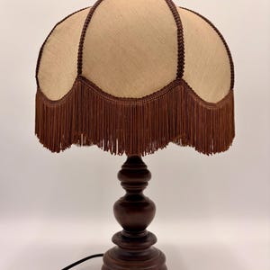 Pu&ograve; includere: Una lampada da tavolo con base in legno tornito marrone scuro e paralume in tessuto beige. Il paralume presenta un bordo smerlato rifinito con un cordoncino intrecciato marrone scuro e lunghe frange marroni abbinate lungo tutto il bordo.