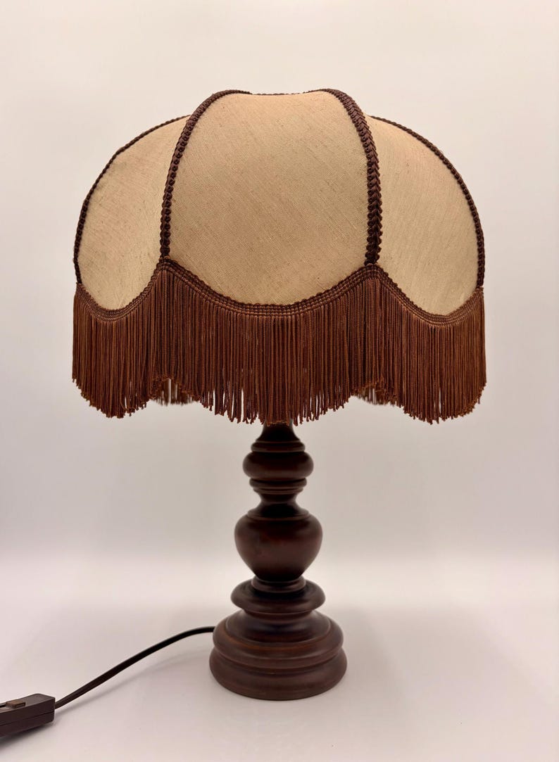 Pu&ograve; includere: Una lampada da tavolo con base in legno tornito marrone scuro e paralume in tessuto beige. Il paralume presenta un bordo smerlato rifinito con un cordoncino intrecciato marrone scuro e lunghe frange marroni abbinate lungo tutto il bordo.