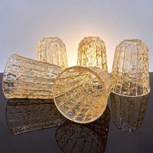 May include: A set of five clear glass light shades featuring a textured, ribbed surface with embedded gold-colored flecks. The shades are cylindrical with a slightly flared opening, displaying a translucent quality that catches the light.