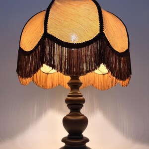 Pu&ograve; includere: Una lampada da tavolo con base in legno marrone scuro dal design tornito e a gradini. La lampada presenta un paralume smerlato color marrone chiaro testurizzato, rifinito con bordi neri e lunghe frange marroni pendenti dal bordo inferiore.