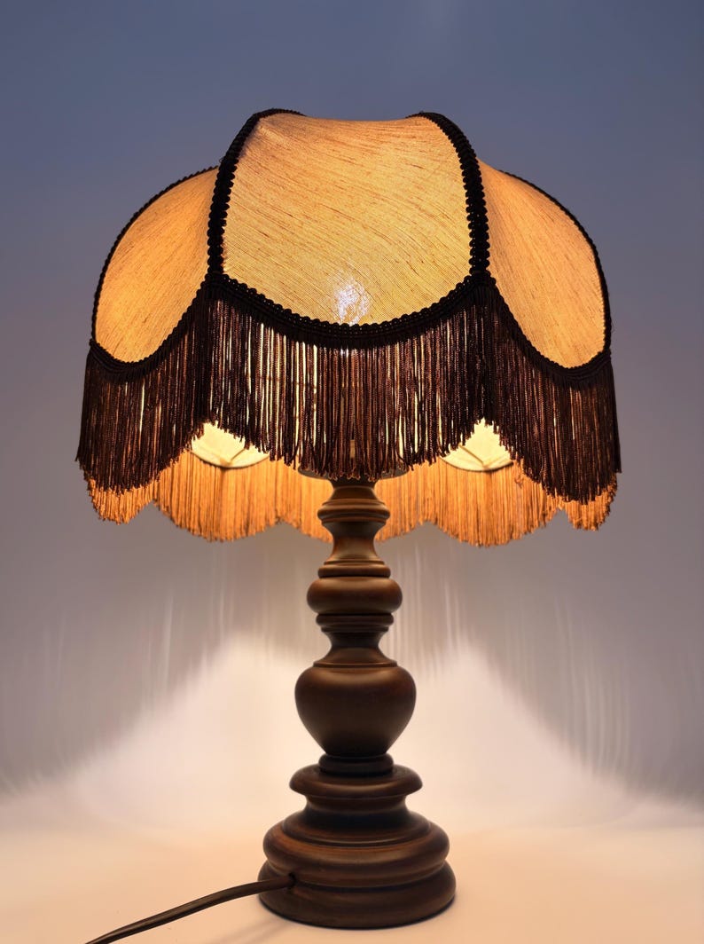 Pu&ograve; includere: Una lampada da tavolo con base in legno marrone scuro dal design tornito e a gradini. La lampada presenta un paralume smerlato color marrone chiaro testurizzato, rifinito con bordi neri e lunghe frange marroni pendenti dal bordo inferiore.