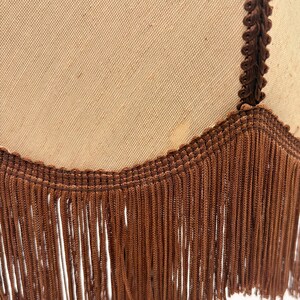 Pu&ograve; includere: Un paralume in tessuto beige con un bordo intrecciato marrone scuro lungo la cucitura verticale e un bordo inferiore smerlato rifinito con lunghe frange marrone scuro che pendono dalla base del paralume.