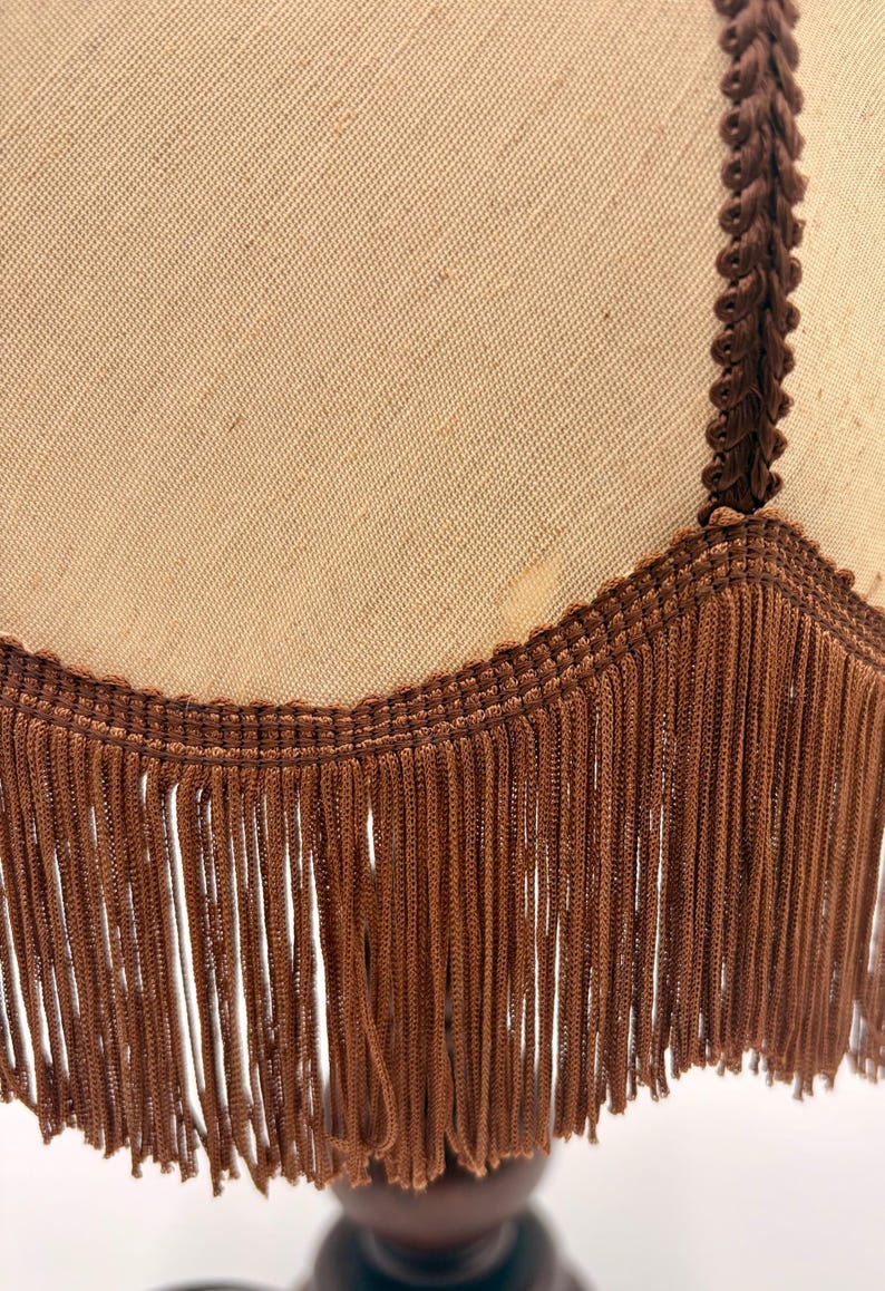 Pu&ograve; includere: Un paralume in tessuto beige con un bordo intrecciato marrone scuro lungo la cucitura verticale e un bordo inferiore smerlato rifinito con lunghe frange marrone scuro che pendono dalla base del paralume.