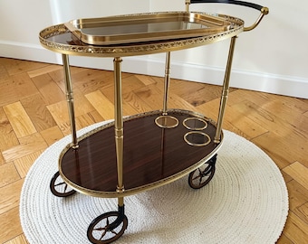 Op de afbeelding: Een ovale, tweelaagse barwagen met een donkere houten afwerking en een goudkleurig metalen frame. De bovenste laag heeft een uitneembaar dienblad. De wagen heeft wielen en een gebogen handvat. De wagen staat op een wit tapijt.