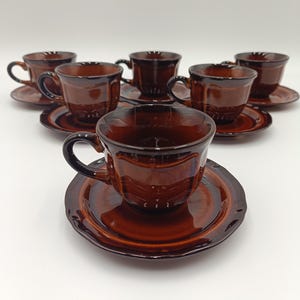 Può includere: Un set di sei tazze da tè e piattini in ceramica marrone. Le tazze hanno una forma arrotondata con un manico e i piattini sono rotondi con un bordo leggermente rialzato. Il set è disposto su una superficie bianca.