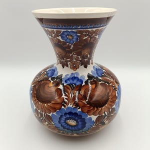 Op de afbeelding: Een keramische vaas met een bolvormige body en uitlopende hals, handbeschilderd met een bloemenmotief. De bloemen zijn voornamelijk bruin en blauw, tegen een witte achtergrond. De rand is bruin en de vaas heeft een glanzende afwerking.