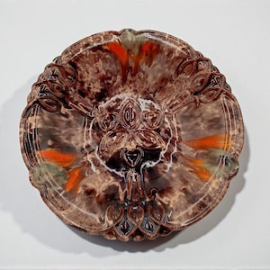 Könnte beinhalten: Ein dekorativer Keramikteller mit einem braunen, orangefarbenen und weißen Marmormuster. Der Teller hat einen detaillierten, verzierten Rand und ein zentrales Blumenmuster. Die Farben verschmelzen zu einem einzigartigen und künstlerischen Aussehen.