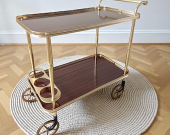 Op de afbeelding: Een serveerwagen met twee niveaus, met een goudkleurig frame en donkere houten planken. De wagen heeft een handvat, decoratieve randen en kleine wielen. De wagen staat op een rond, geweven tapijt.
