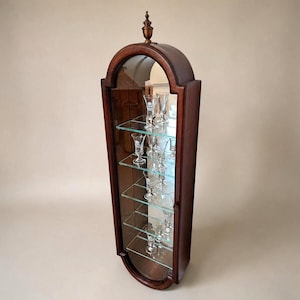 Peut inclure: Une vitrine murale haute et arquée en bois brun foncé avec une porte vitrée et un panneau arrière en miroir. Elle comporte cinq étagères horizontales en verre contenant plusieurs petits verres à pied transparents à l'intérieur.