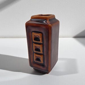 Può includere: Un vaso rettangolare in ceramica con una smaltatura marrone scuro lucida. Il vaso presenta una fila verticale di tre rientranze quadrate sulla parte anteriore, un'apertura superiore piatta e una finitura superficiale liscia e riflettente.