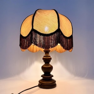 Pu&ograve; includere: Una lampada da tavolo con base in legno marrone scuro e paralume testurizzato di colore chiaro. Il paralume presenta un bordo smerlato rifinito con frange marrone scuro e profili dello stesso colore lungo le cuciture, creando un aspetto stratificato.