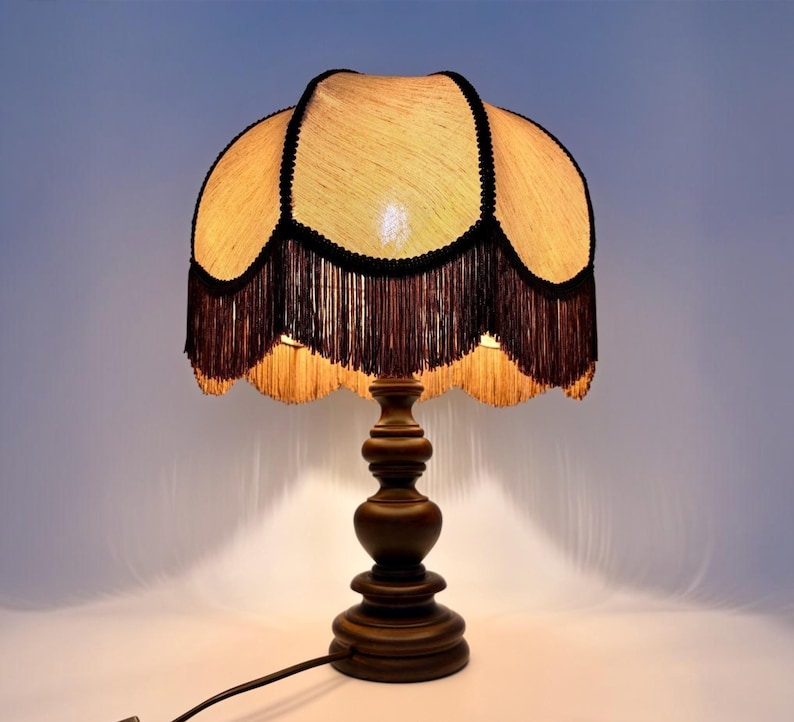 Pu&ograve; includere: Una lampada da tavolo con base in legno marrone scuro e paralume testurizzato di colore chiaro. Il paralume presenta un bordo smerlato rifinito con frange marrone scuro e profili dello stesso colore lungo le cuciture, creando un aspetto stratificato.