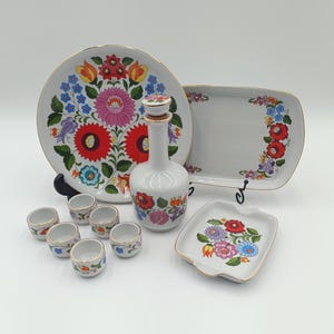 Op de afbeelding: Een witte keramische serveerset met een bloemmotief. De set bevat een groot rond bord, een rechthoekige schaal, een klein vierkant schaaltje, een fles en diverse kleine kopjes. Het bloemmotief toont rode, roze, blauwe en gele bloemen met een gouden rand.