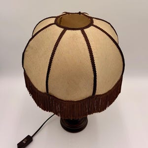 Pu&ograve; includere: Un paralume in tessuto beige dalla forma smerlata, caratterizzato da un bordo intrecciato marrone scuro lungo le cuciture e da una lunga frangia marrone scuro sul bordo inferiore. Il paralume &egrave; fissato su una base marrone scuro con cavo nero.