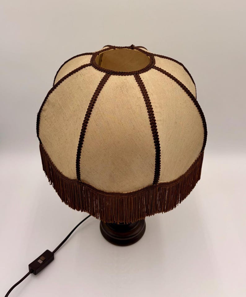 Pu&ograve; includere: Un paralume in tessuto beige dalla forma smerlata, caratterizzato da un bordo intrecciato marrone scuro lungo le cuciture e da una lunga frangia marrone scuro sul bordo inferiore. Il paralume &egrave; fissato su una base marrone scuro con cavo nero.