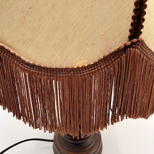 Pu&ograve; includere: Una lampada da tavolo con paralume in tessuto beige dal bordo inferiore smerlato. Il paralume &egrave; rifinito con un cordoncino intrecciato marrone scuro e una lunga frangia densa dello stesso colore. La base &egrave; in materiale marrone scuro e arrotondata.