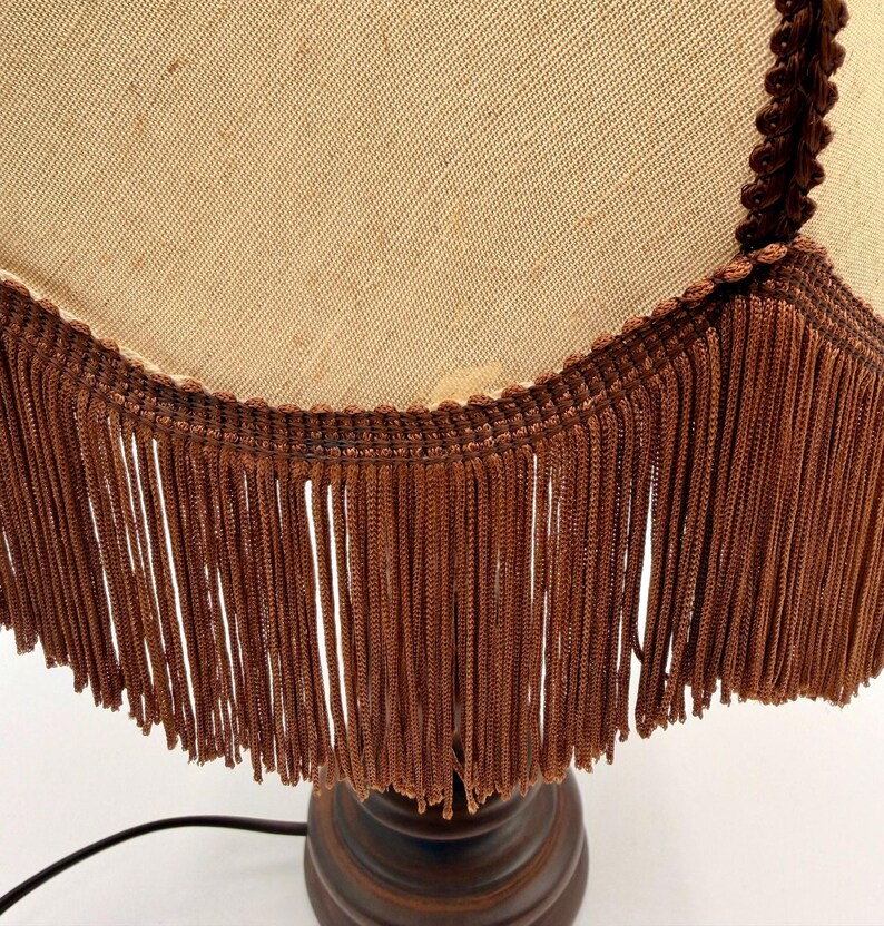 Pu&ograve; includere: Una lampada da tavolo con paralume in tessuto beige dal bordo inferiore smerlato. Il paralume &egrave; rifinito con un cordoncino intrecciato marrone scuro e una lunga frangia densa dello stesso colore. La base &egrave; in materiale marrone scuro e arrotondata.