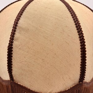Pu&ograve; includere: Un paralume a forma di cupola con una superficie in tessuto beige chiaro divisa in segmenti da un bordo intrecciato marrone scuro. Il bordo inferiore del paralume &egrave; rifinito con una lunga frangia marrone scuro che pende verticalmente.