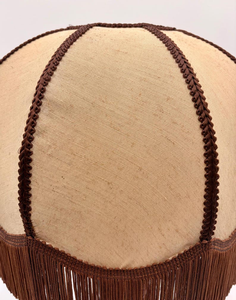 Pu&ograve; includere: Un paralume a forma di cupola con una superficie in tessuto beige chiaro divisa in segmenti da un bordo intrecciato marrone scuro. Il bordo inferiore del paralume &egrave; rifinito con una lunga frangia marrone scuro che pende verticalmente.