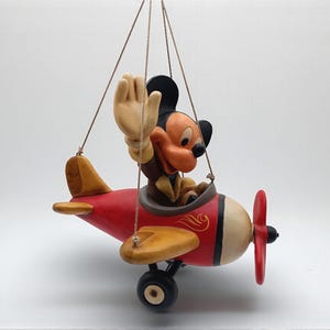 Peut inclure: Figurine vintage de Mickey Mouse dans un avion rouge, jaune et crème. Mickey salue depuis le cockpit. L'avion a une hélice rouge et des roues noires. La figurine est suspendue à une ficelle.