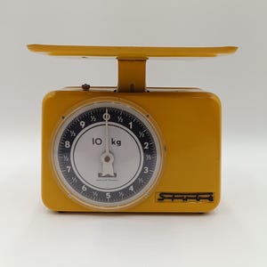 Op de afbeelding: Een gele vintage keukenweegschaal met een ronde wijzerplaat die metingen tot 10 kg weergeeft. De weegschaal heeft een rechthoekige basis, een platte bovenkant en een zwart-witte wijzerplaat met duidelijke markeringen. De merknaam is zichtbaar.