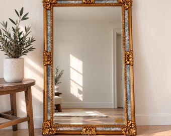 May include: A large rectangular floor mirror featuring an ornate gold frame with intricate carved detailing at the corners and center. The inner border of the frame has a blue mottled finish. The mirror reflects a room with a wooden floor and a doorway.