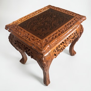Op de afbeelding: Een rechthoekige, bruine houten salontafel met ingewikkelde bloemsnijwerken. De tafel heeft een glazen blad en gebogen poten. De gedetailleerde snijwerken zijn zichtbaar op het frame en de poten, met een traditioneel ontwerp. Ongeveer 90 cm lang.