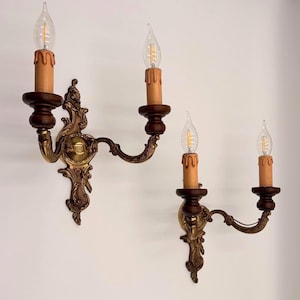 Könnte beinhalten: Zwei antike Wandleuchter mit Vintage-Design. Jeder Leuchter verfügt über einen dunkelbraunen Holzsockel, eine kerzenförmige Glühbirne und einen dekorativen Messingarm. Die Leuchter sind an einer weißen Wand montiert und erzeugen einen klassischen und eleganten Look.
