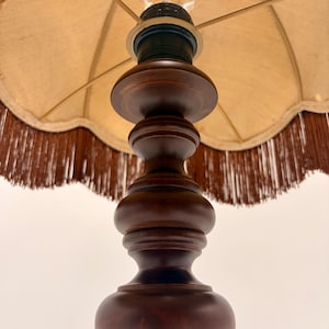 Pu&ograve; includere: Una lampada da tavolo con base in legno tornito marrone scuro dalla silhouette liscia e arrotondata. La lampada &egrave; dotata di un paralume in tessuto beige con bordo smerlato e una lunga frangia marrone scuro che pende dal bordo inferiore.