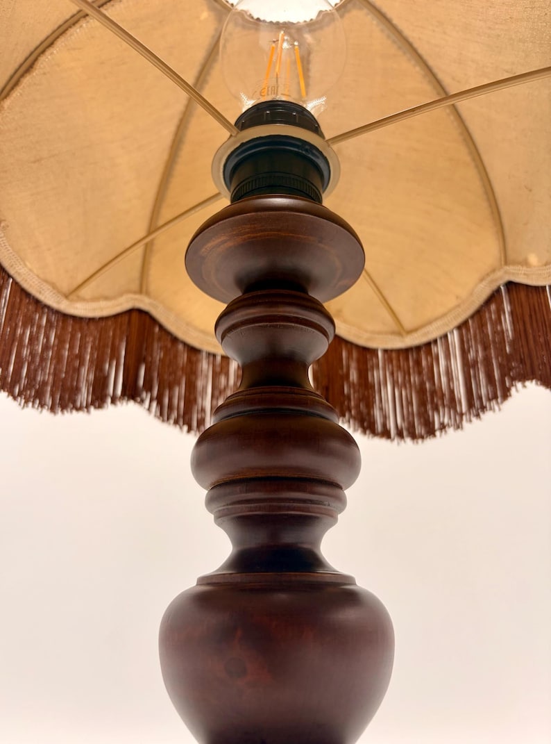 Pu&ograve; includere: Una lampada da tavolo con base in legno tornito marrone scuro dalla silhouette liscia e arrotondata. La lampada &egrave; dotata di un paralume in tessuto beige con bordo smerlato e una lunga frangia marrone scuro che pende dal bordo inferiore.