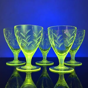Könnte beinhalten: Ein Set aus fünf durchscheinenden gelb-grünen Glasbechern, die jeweils mit einem zarten, geätzten Blattmuster um die Kuppa versehen sind. Die Gläser haben einen kurzen, stabilen Stiel und einen runden Fuß und leuchten in einem kräftigen Farbton.