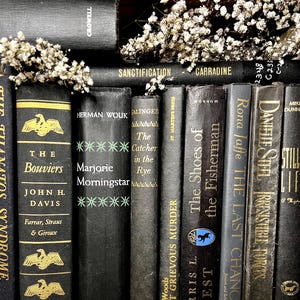 May include: A selection of antique books with black covers and gold lettering. Titles include "The Bouviers," "Marjorie Morningstar," and "The Catcher in the Rye." Small white flowers are arranged amongst the books, creating a classic look.