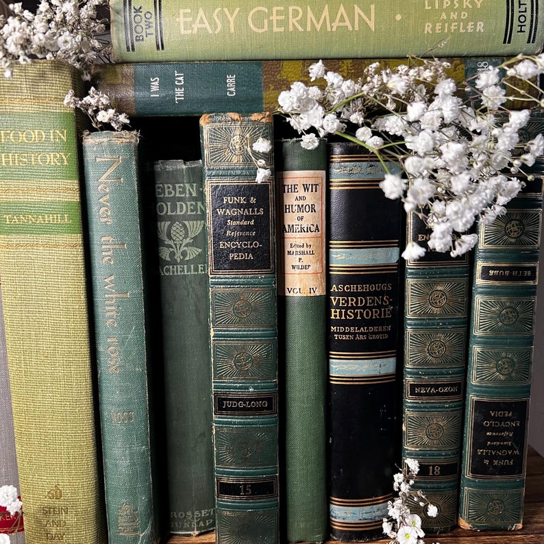 May include: A collection of antique books in various shades of green and gold, with titles like "Easy German" and "Food in History." White flowers are nestled among the spines. The books are arranged vertically, showcasing their aged covers and decorative details.