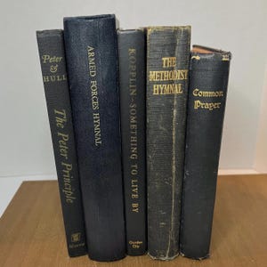 May include: Five antique books with dark covers are displayed. The titles include "The Peter Principle," "Armed Forces Hymnal," "Kopplin - Something to Live By," "The Methodist Hymnal," and "Common Prayer." The text is in gold lettering.